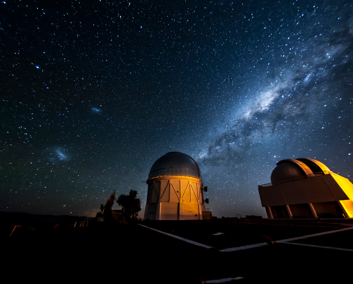 two dimly lit observatories sit as stunted domes beneath a vibrant night sky and a stretch of blue hued milky way.