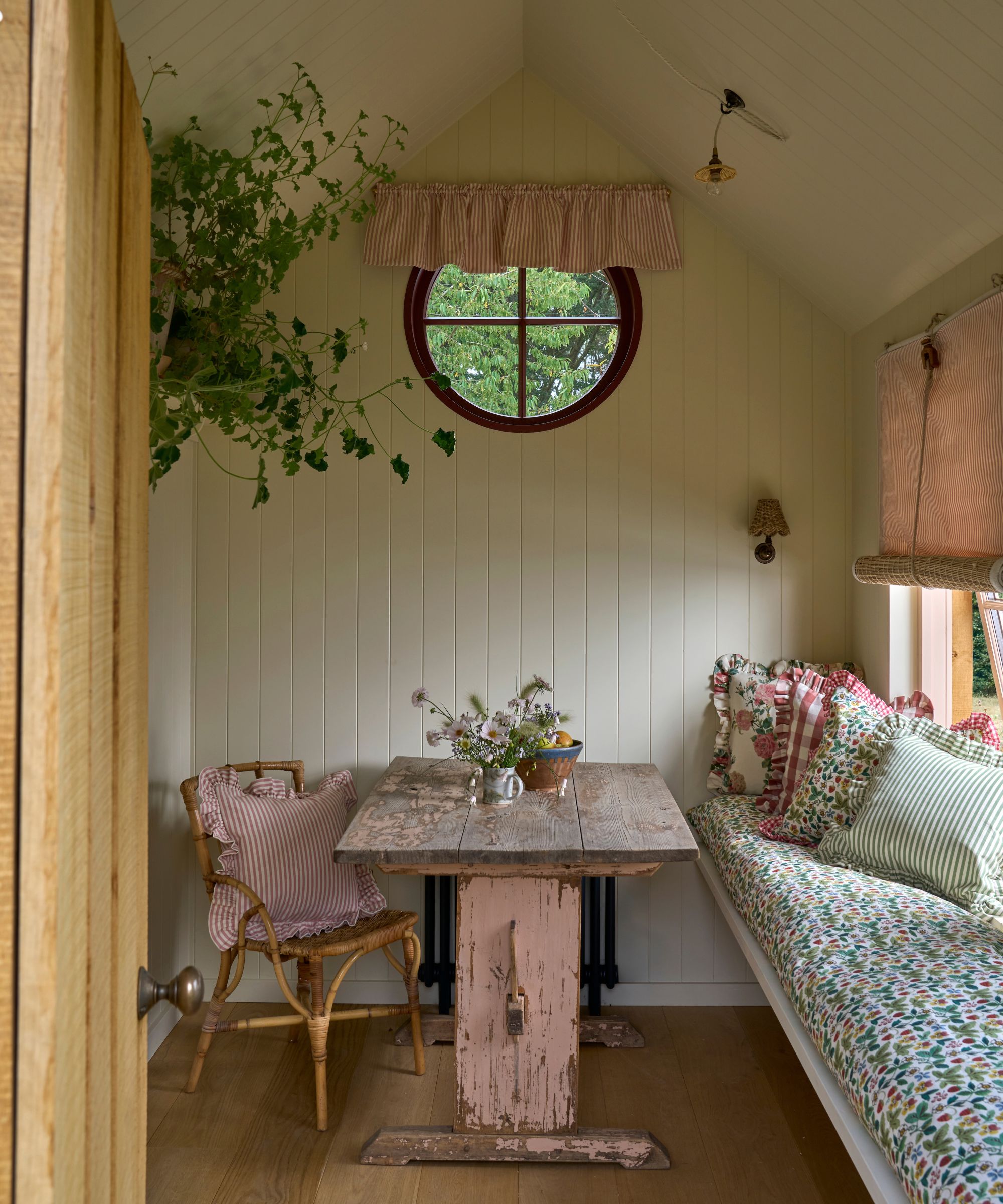 Rustic sunroom with cream panelling walls, a rustic pink dining table and a bench upholstered in floral fabrics