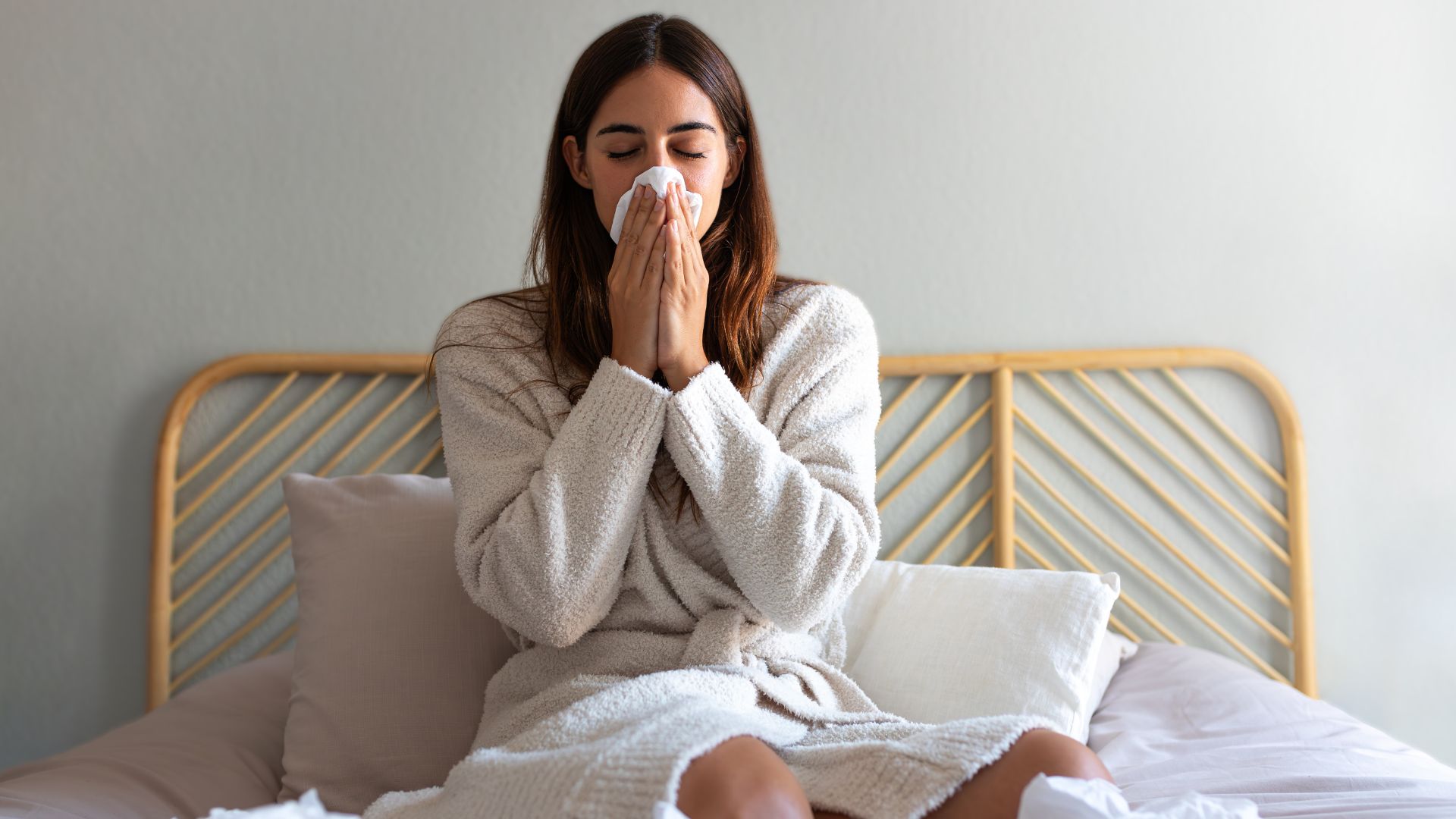A woman sits in bed wearing a dressing gown and blowing her nose.