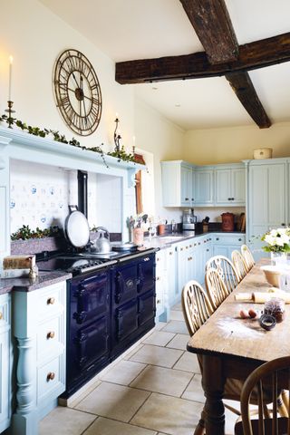 A country kitchen with light blue cabinets with wood dining table, large range cooker and original beams