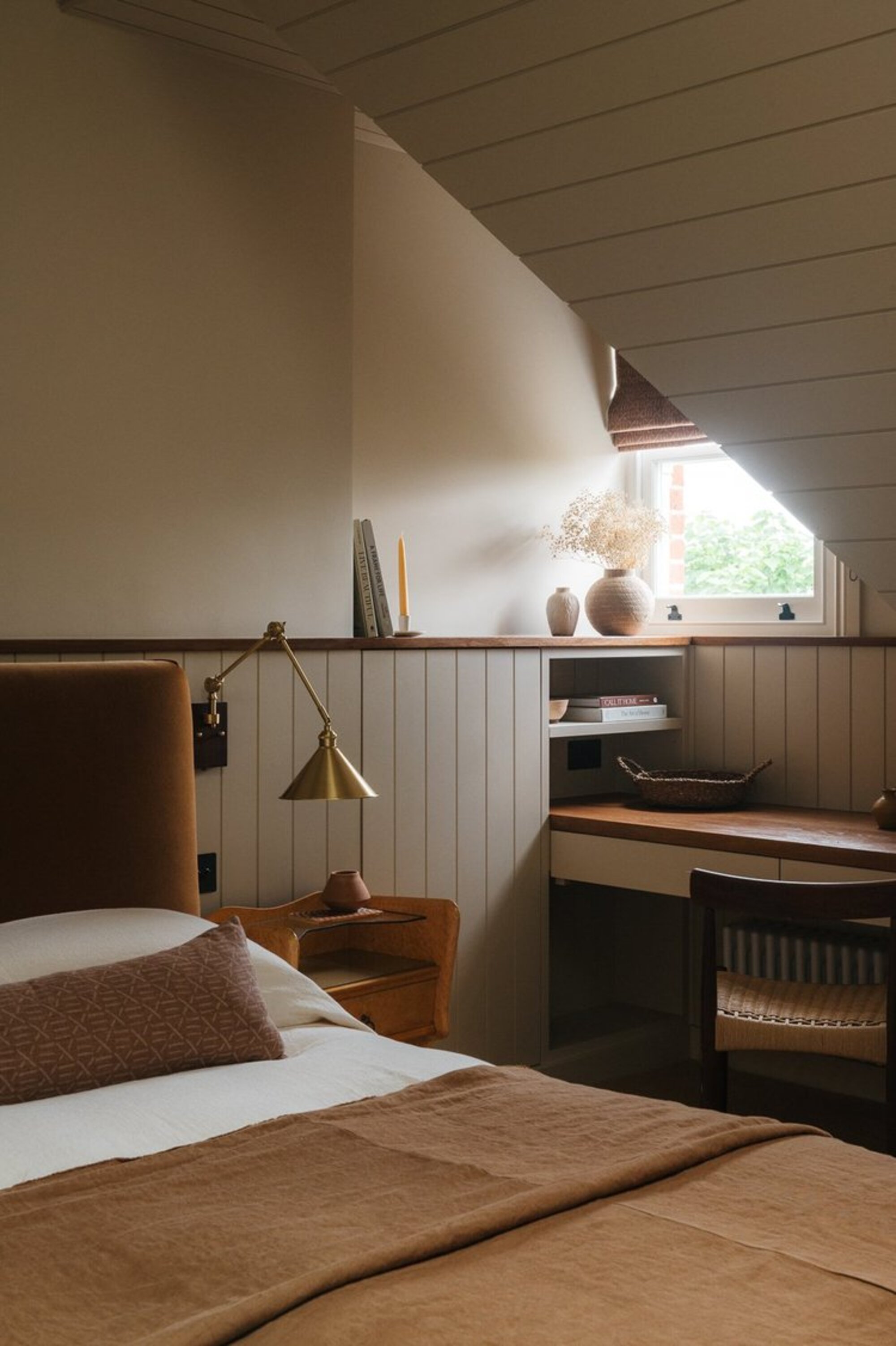 modern bedroom with sloped ceiling and window in nook with wall paneling and built-in desk with bed made with white sheets and orange blanket