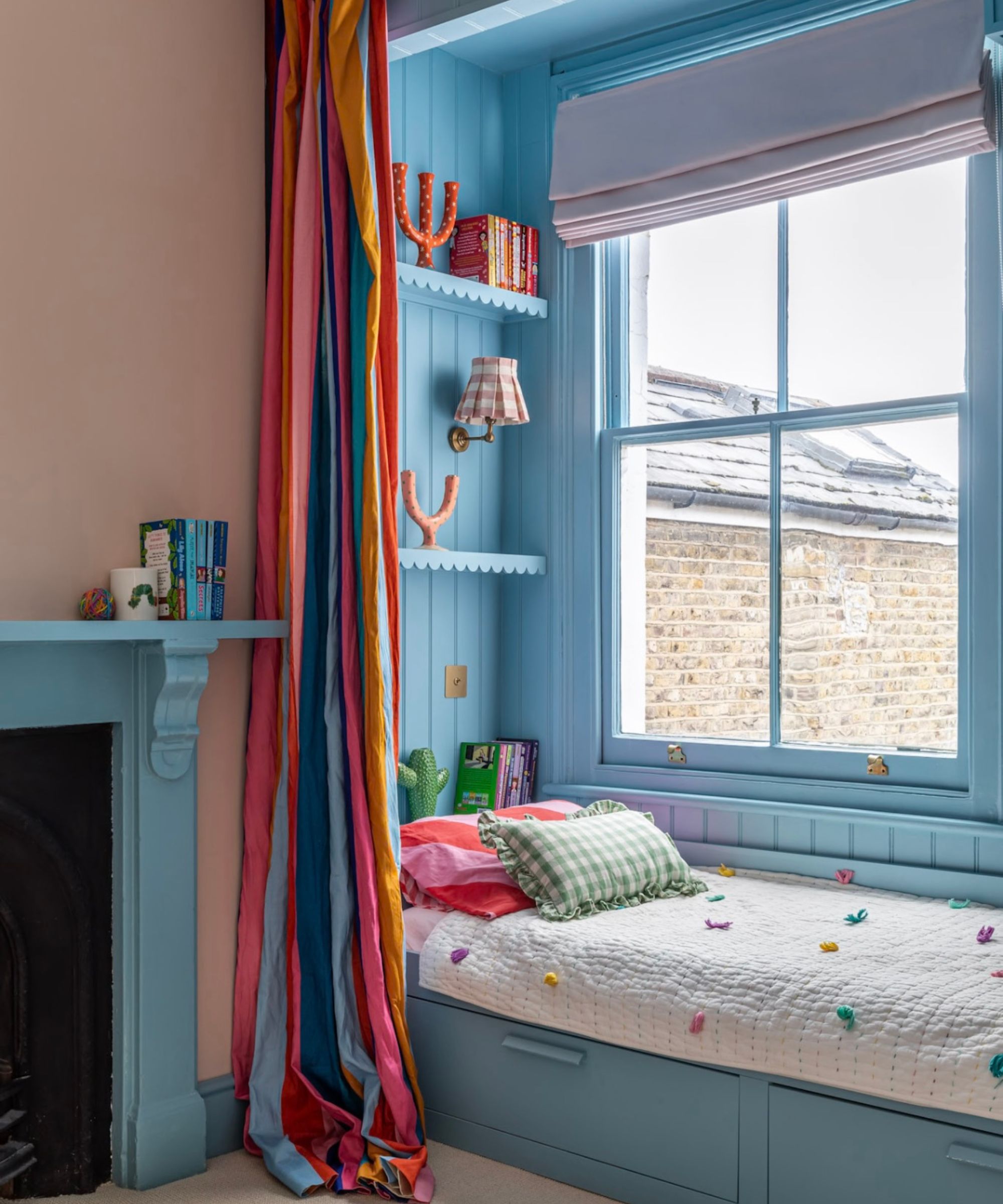 A child's bedroom with plaster pink walls, a blue cabin bed beside a window with a colorful curtain, and a blue fireplace.