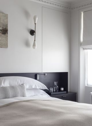 white bedroom with dark gray wood headboard and combined side table, oatmeal blanket, white bedding, wall light, artwork