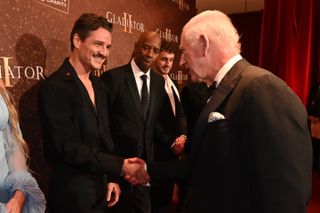 Pedro Pascal wears a black suit with no shirt underneath while he shakes hands with King Charles, who is wearing a tuxedo