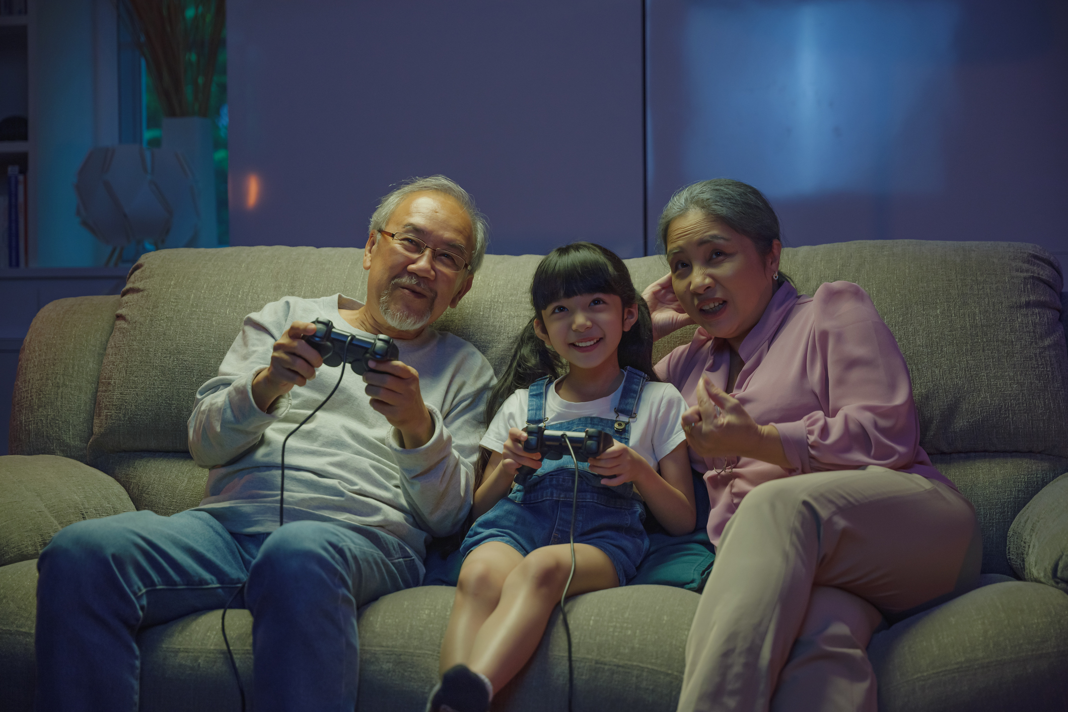 Happy Asian family bonding over a video game on a cozy couch, little girl playing video games console with senior grandparents together with controller joysticks while sitting on the sofa at night