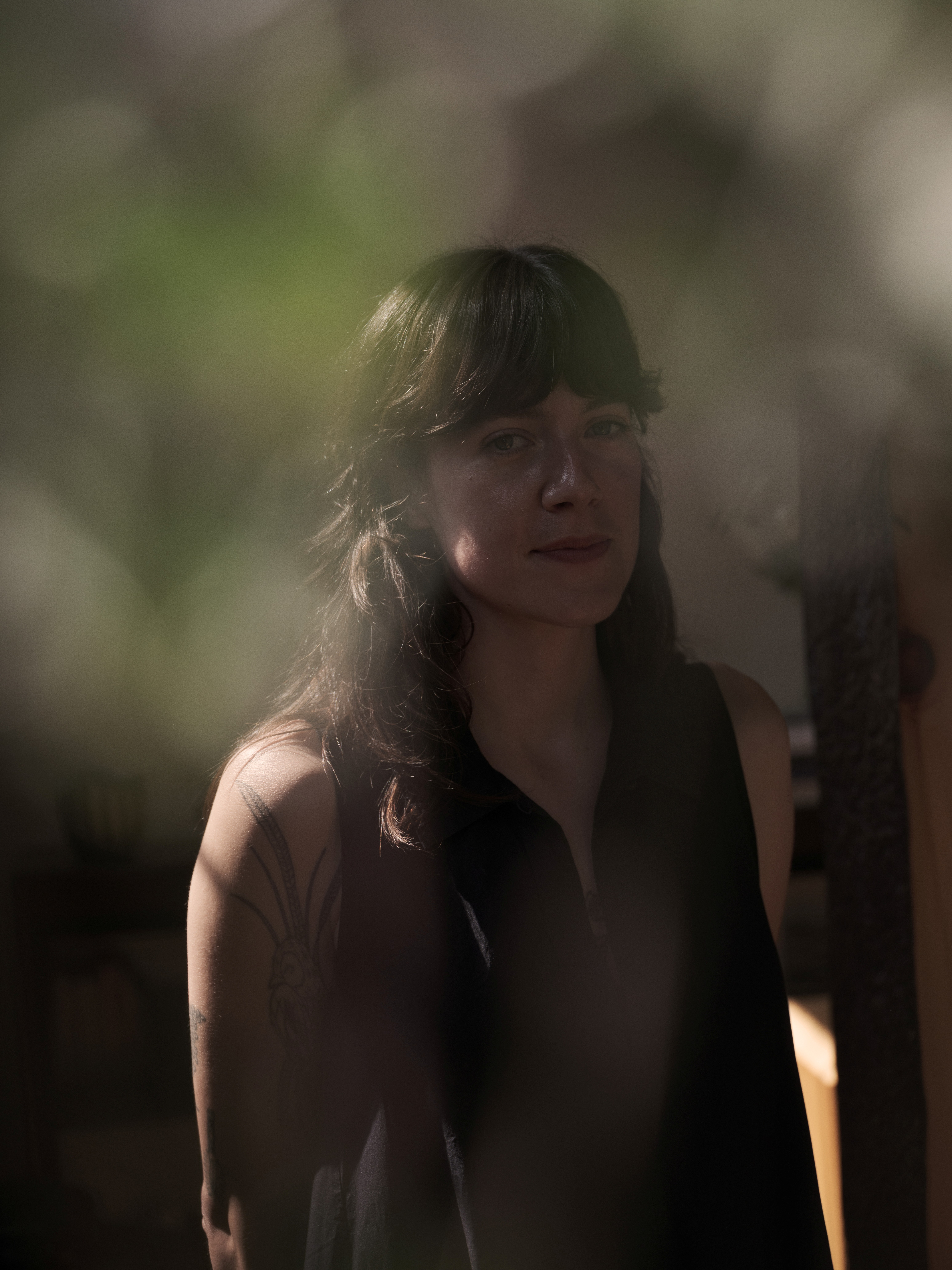 A young woman with dark hair and bangs wearing a brown top in a bokeh-style portrait surrounded by greenery.
