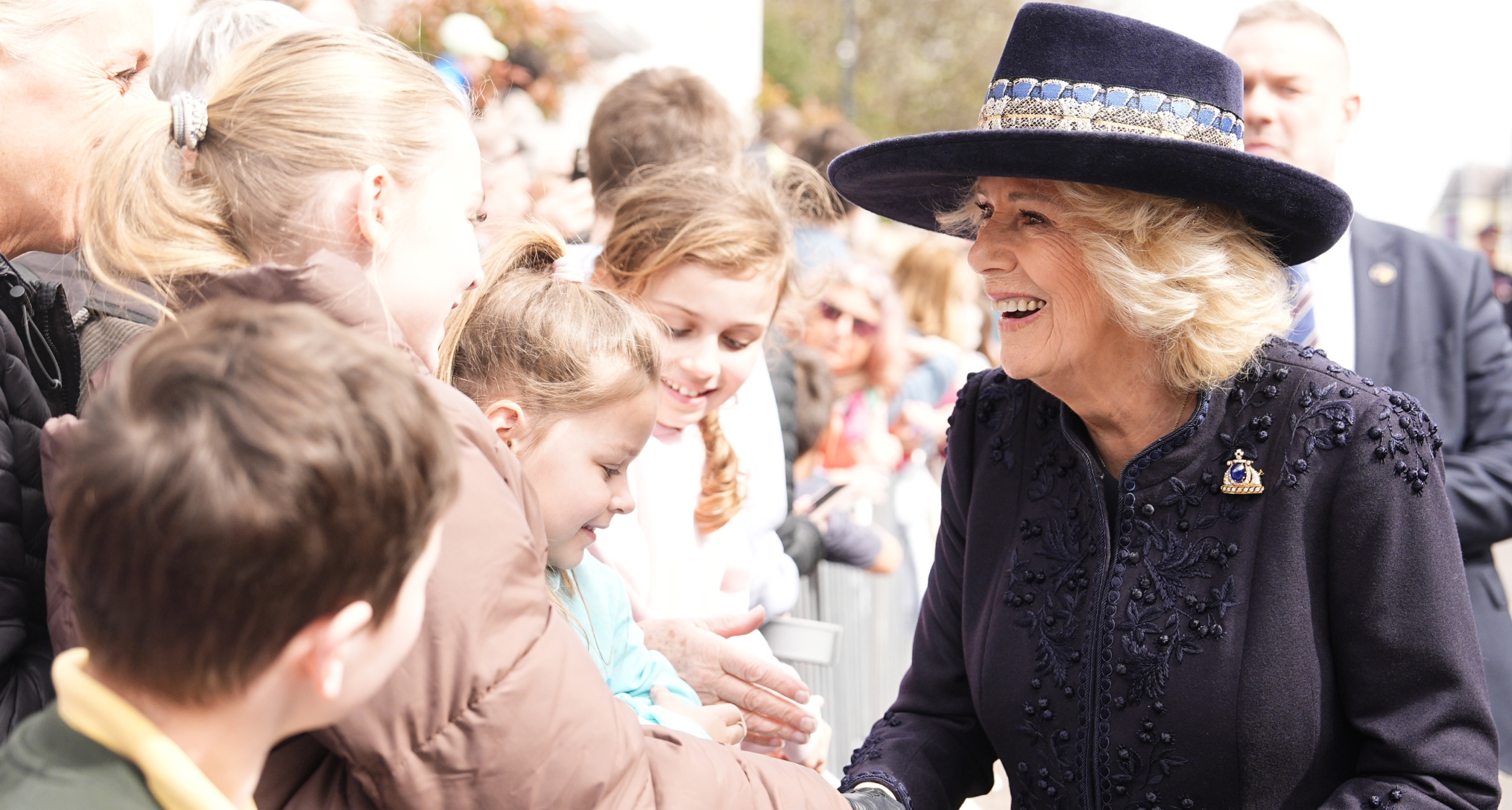 Queen Camilla wearing a blue coat and hat shaking hands with people
