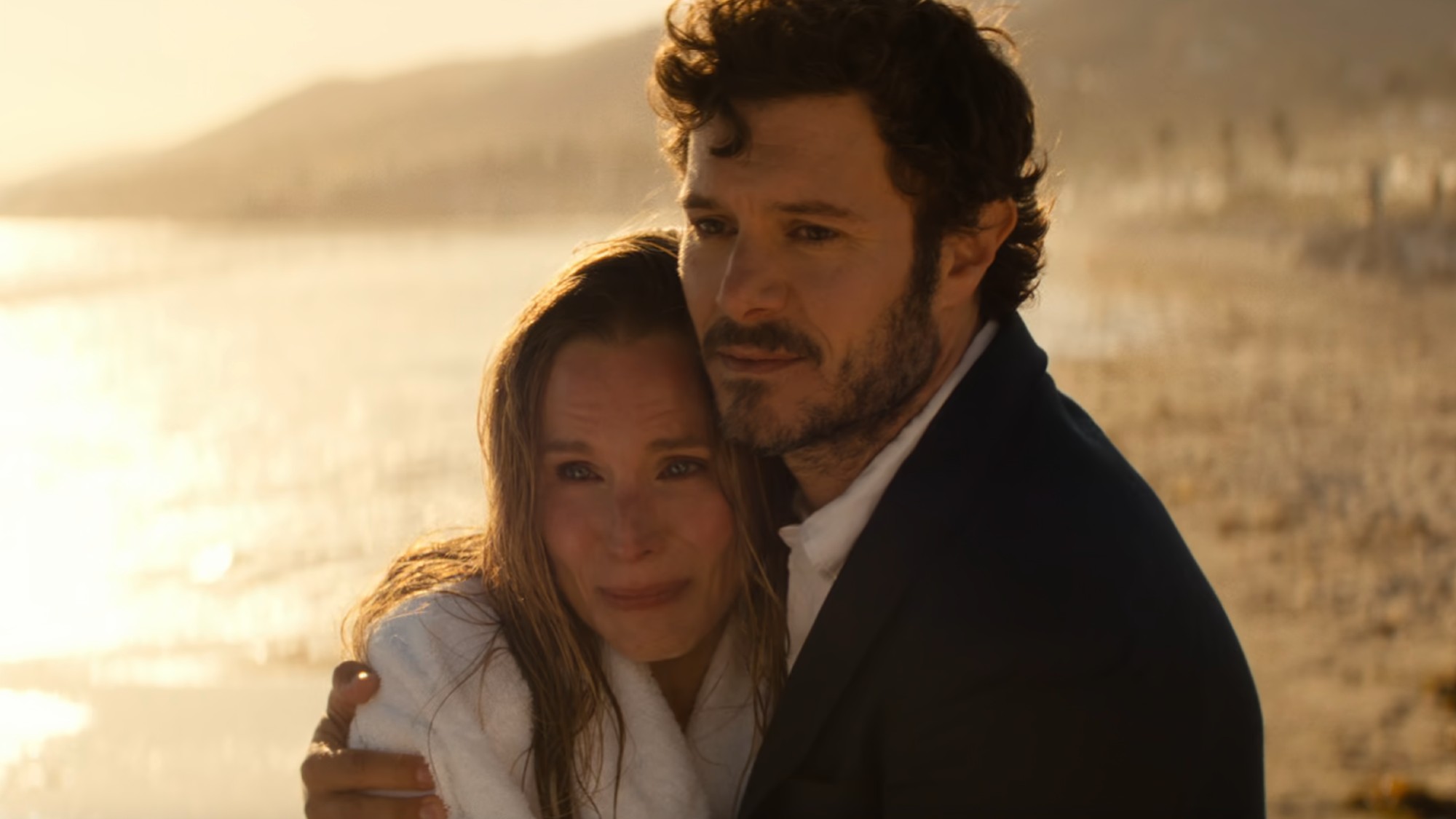 From left to right: Kristen Bell as Joanne and Adam Brody as Noah standing on a beach and hugging each other while looking forward in Nobody Wants This. 