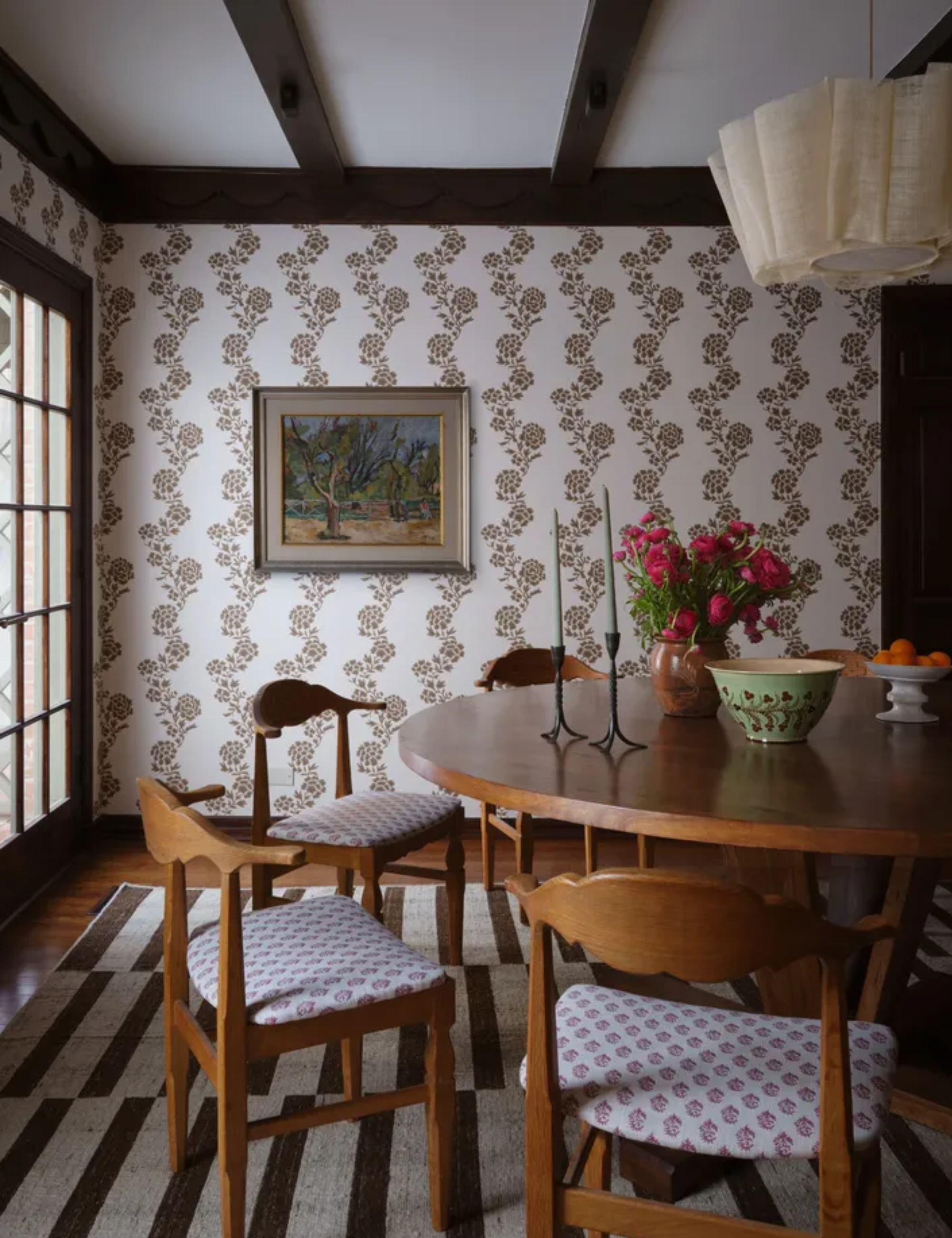 A vertical shot of a wooden dining set on a striped rug, featuring chairs with pink patterned cushions and walls adorned with brown floral wallpaper
