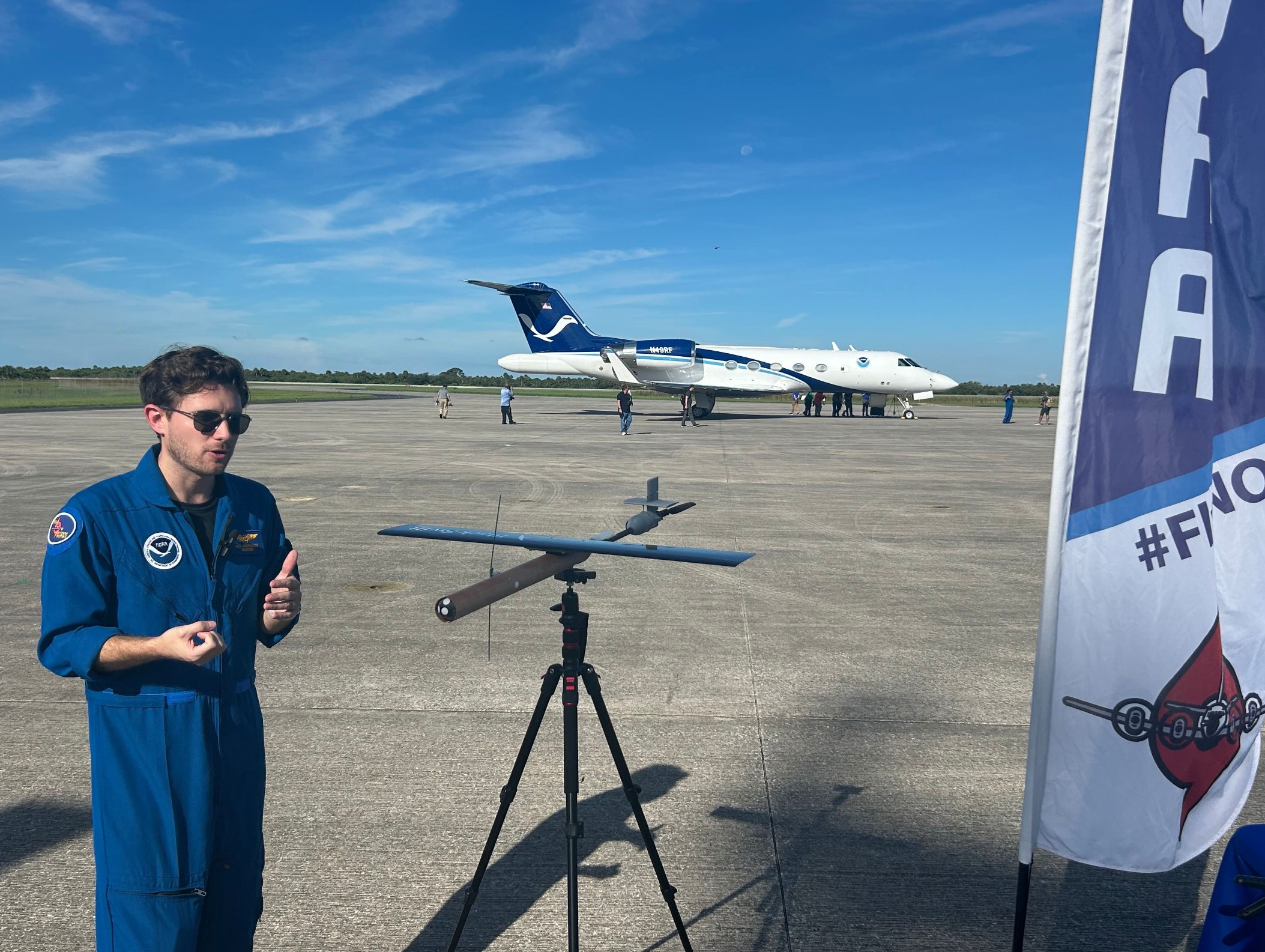 an aviator in a blue flight suit stands beside a small drone with a large white jet in the background