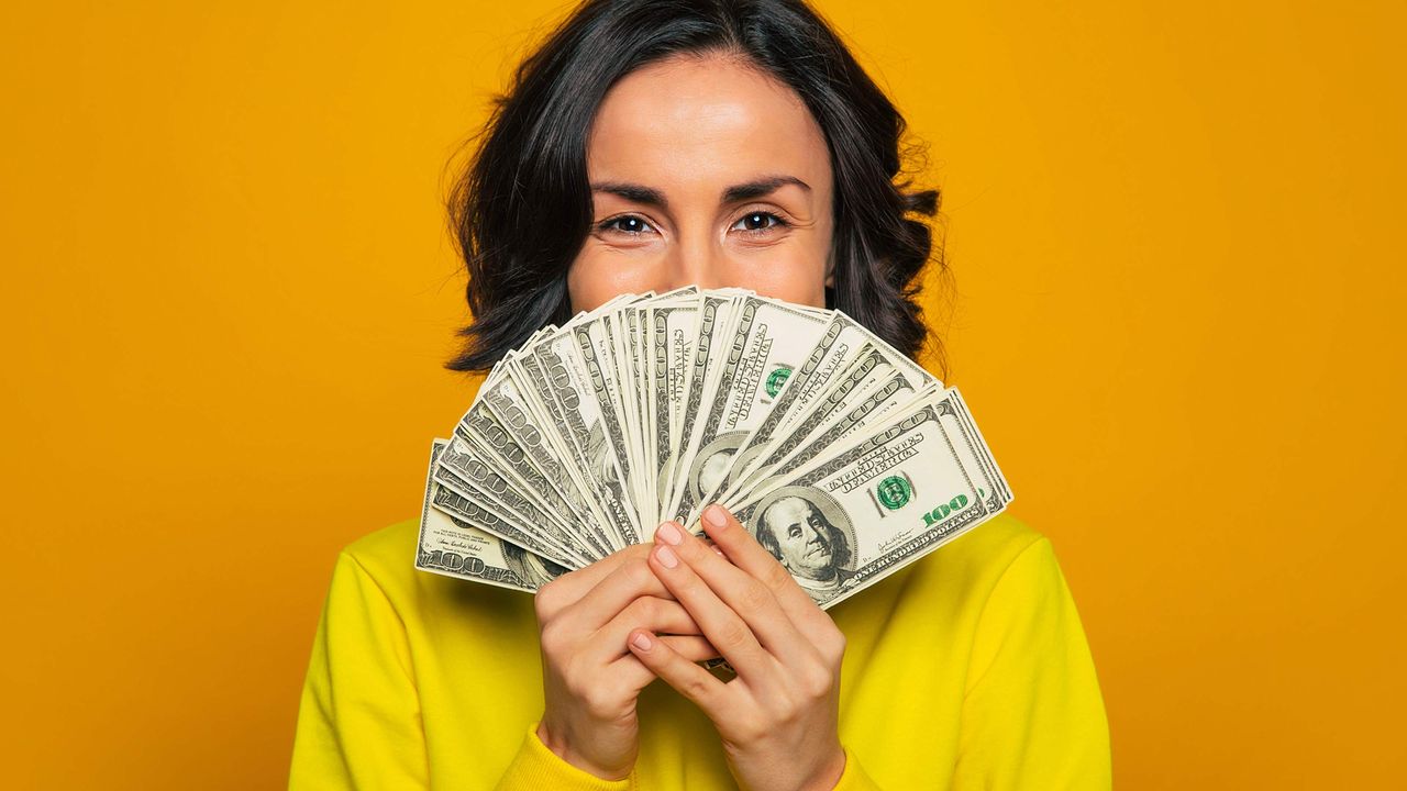 A woman holds a bunch of money like a fan in front of the bottom half of her face.