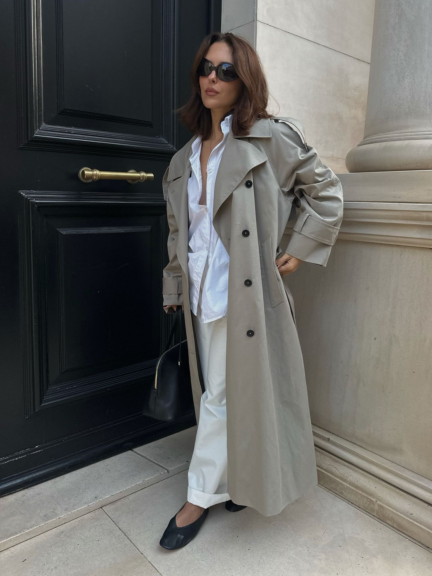 Style influencer Debora Rosa poses in a long gray trench coat, white button-down shirt, ivory cuffed pants, a black bag, and black v-cut flats