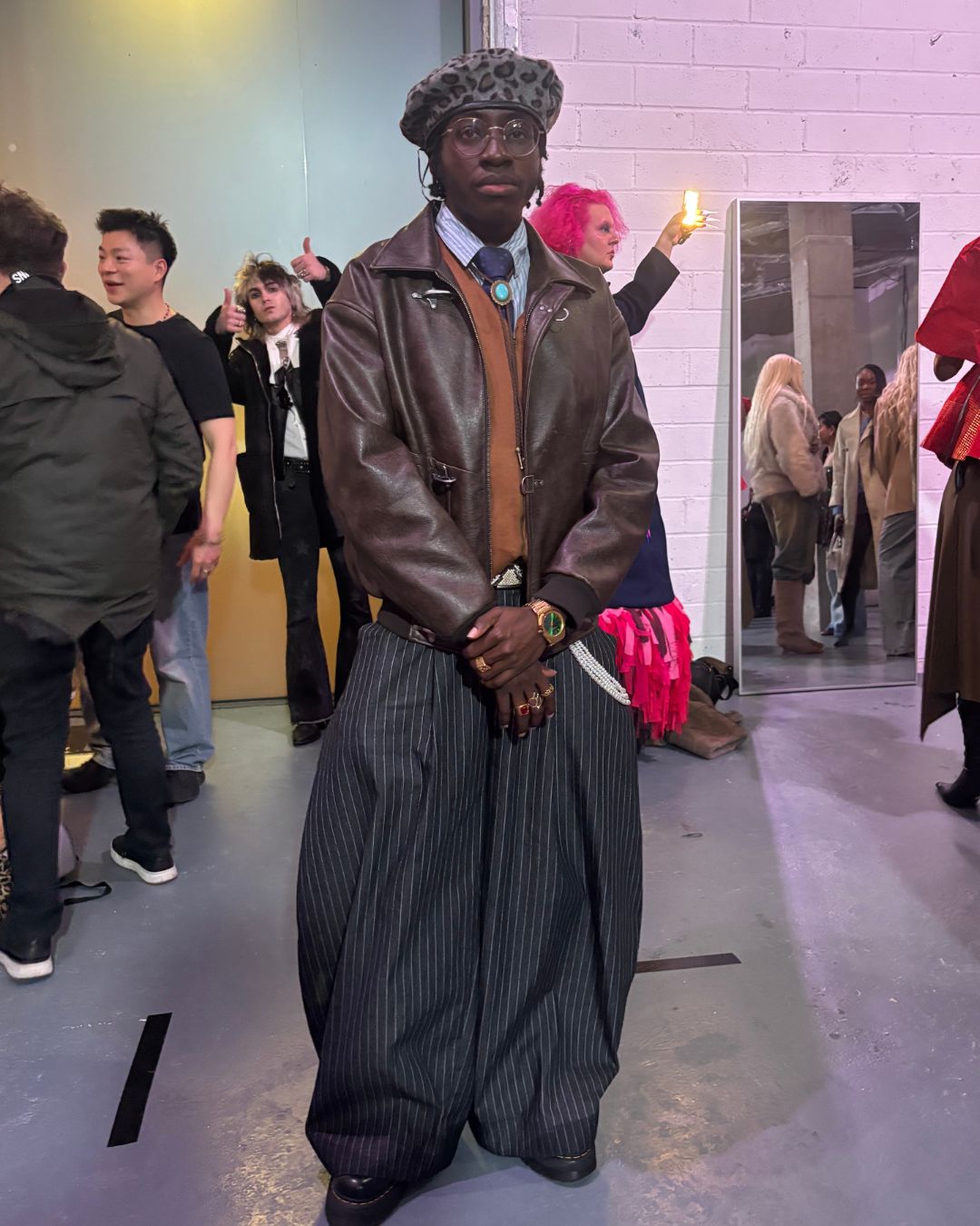 an image of Timmy &amp;ndash; an fashion influencer (@timzino._) in his outfit for London Fashion Week, featuring a leopard print beret, a layered brown leather jacket, and super wide leg / barrel leg pinstripe trousers