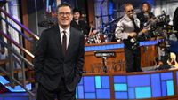 Stephen Colbert stands in front of The Late Show band during a final episode on CBS. He has on a suit and tie.