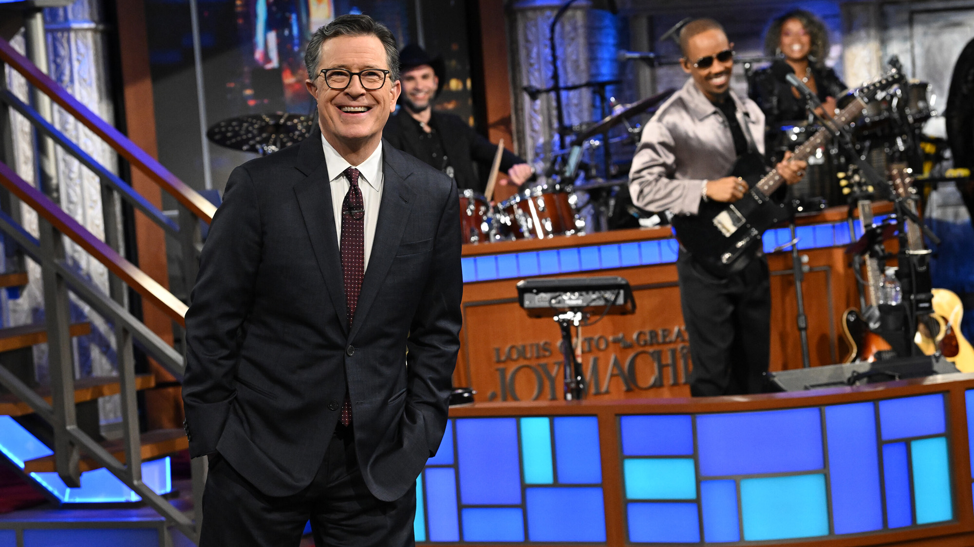 Stephen Colbert stands in front of The Late Show band during a final episode on CBS. He has on a suit and tie.