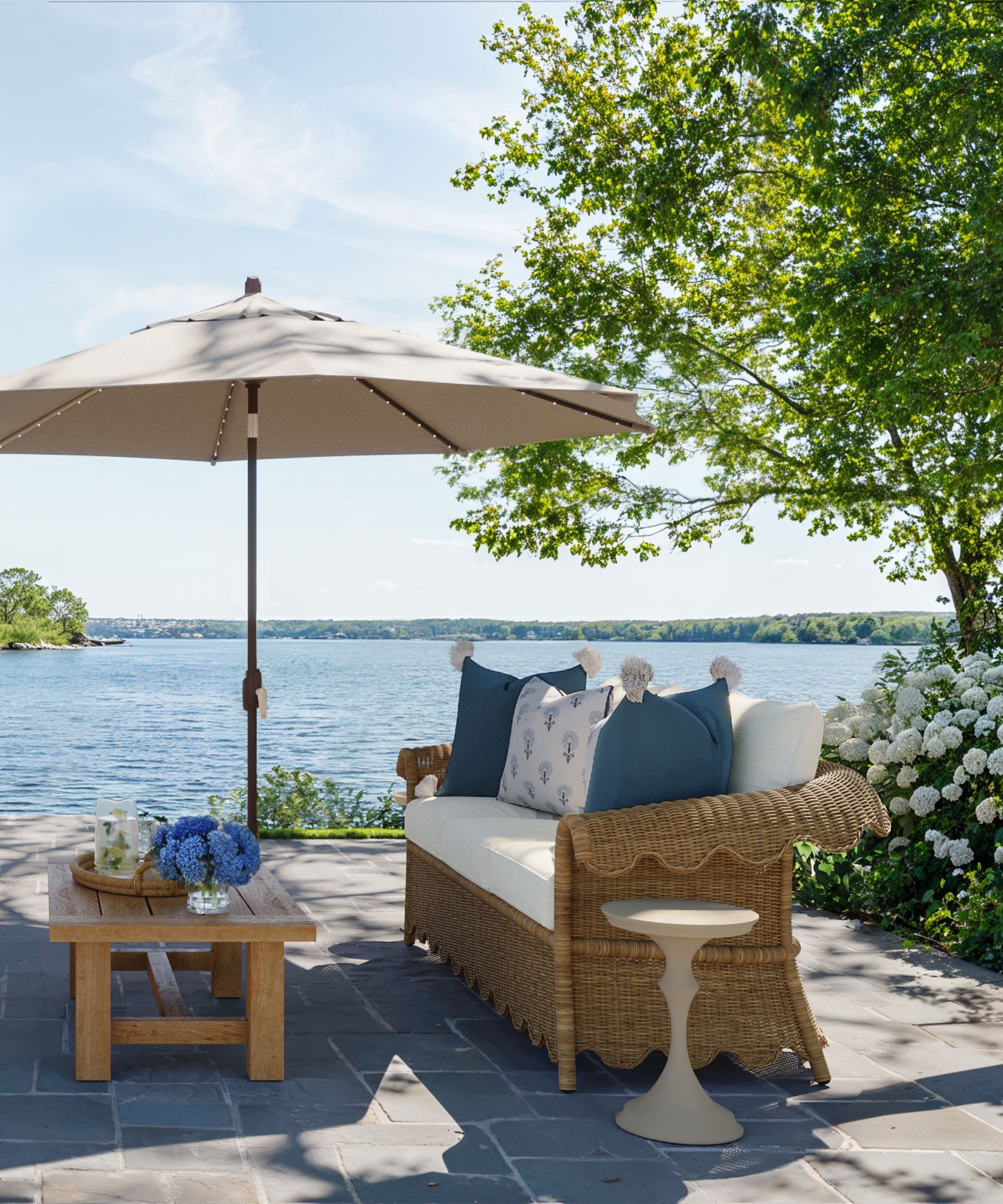 lakeside patio with large hydrangea bushes, a teak coffee table, white patio umbrella, and rattan scalloped outdoor furniture with blue and white cushions