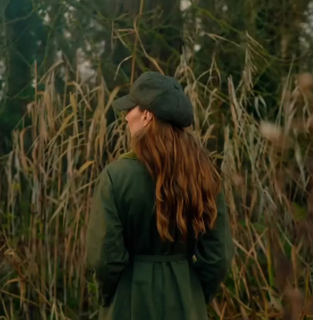 Kate Middleton standing in front of tall grass in a green coat and hat with her back to the camera