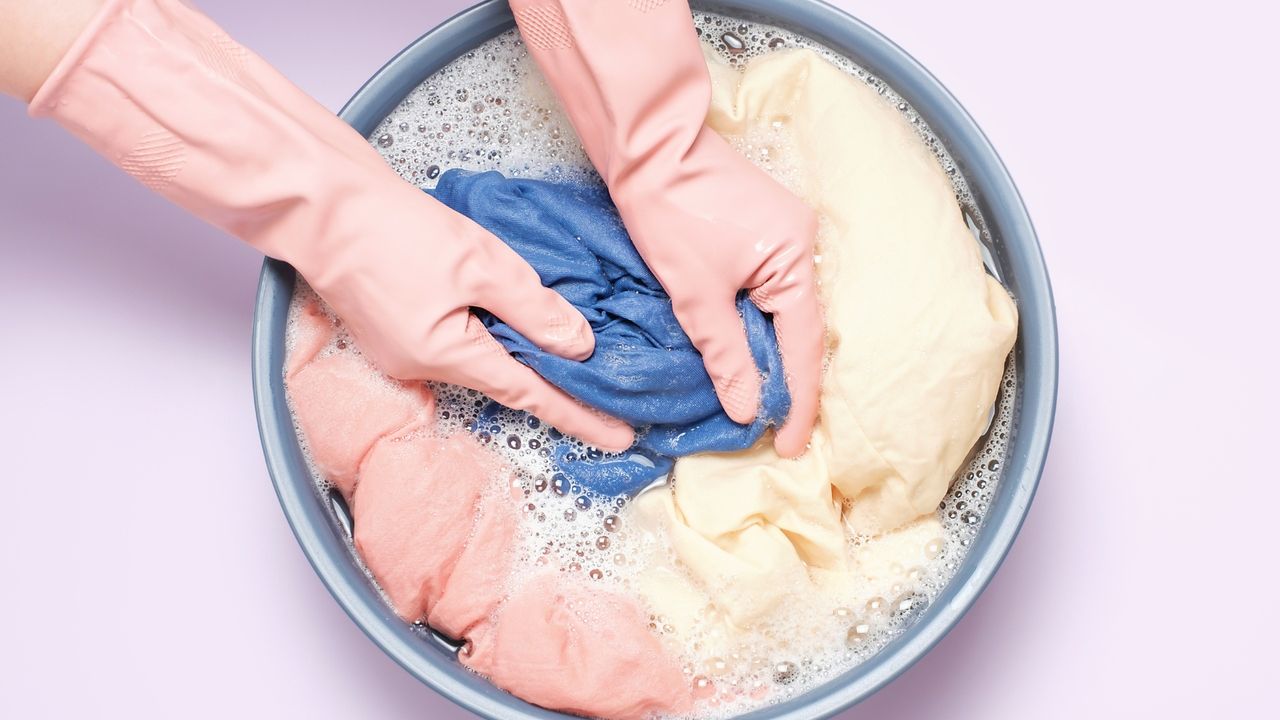 Hands in rubber gloves, handwashing laundry in basin on lilac background, top view