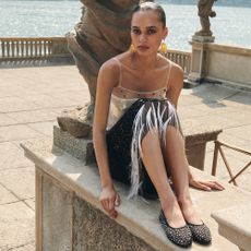 Model sat on a low wall next to a lake wearing a sequin Tory Burch party dress, big gold earrings and studded ballet flats sold for Tory Burch discount codes