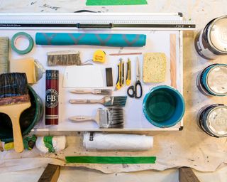 The tools you need to learn how to wallpaper a room, including paintbrushes, rollers, a tape measure, stanley knives and paste.
