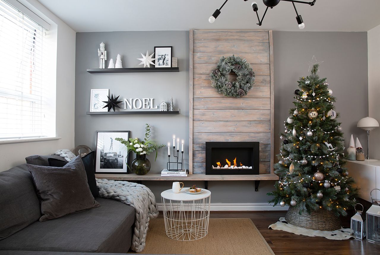 Grey sofa with wooden panelled fireplace, Christmas tree, brown weave rug and black shelving with &#039;Noel&#039; letter decorations and a pine cone wreath