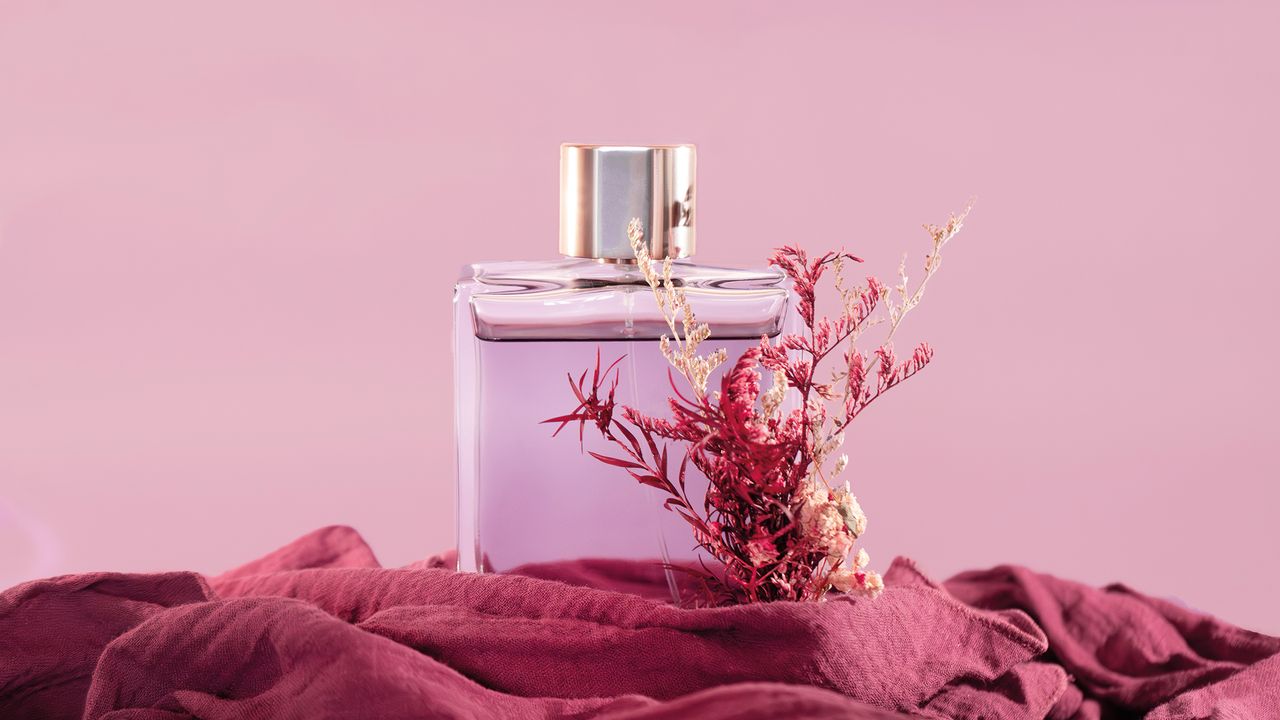 A clear glass perfume bottle with a gold cap sits on a burgundy fabric, adorned with delicate dried flowers, against a soft pink background