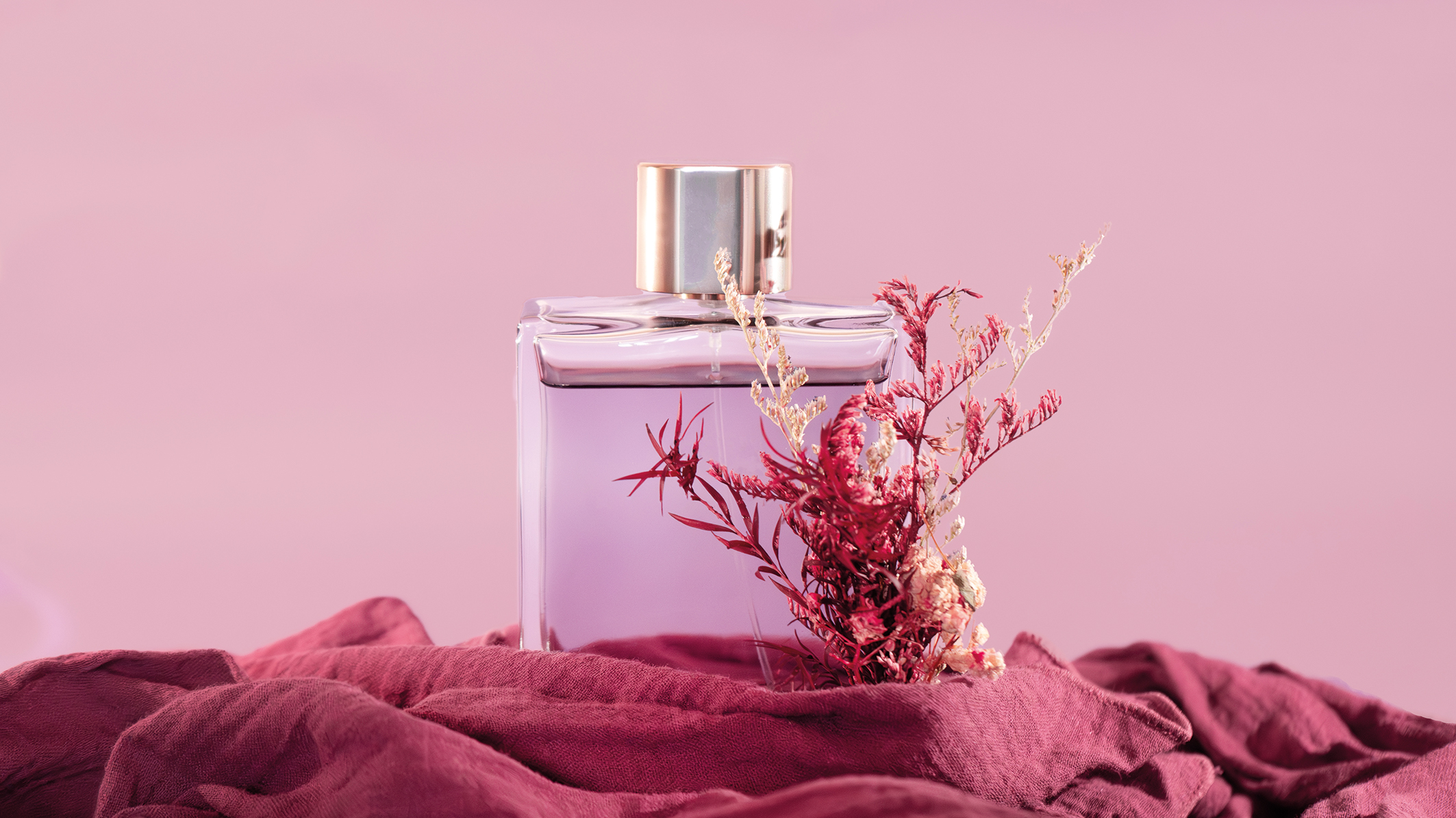 A clear glass perfume bottle with a gold cap sits on a burgundy fabric, adorned with delicate dried flowers, against a soft pink background