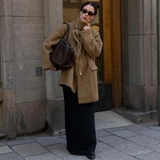 annabel wears black sunglasses a tan coat with a scarf, brown leather bag, and black maxi skirt.