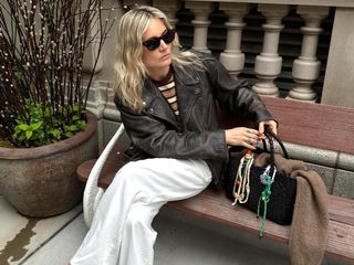 British influencer Lucy Williams sitting on a bench in NYC wearing cat eye sunglasses, a striped tee, brown leather moto jacket, white pants, and an embellished woven leather bag