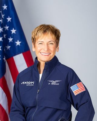 headshot of peggy whitson smiling wearing an Axiom space blue jacket with the American flag on the left arm and a large American flag behind her.l