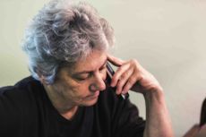 An impatient older woman is waiting on hold on the telephone while sitting at her desk.