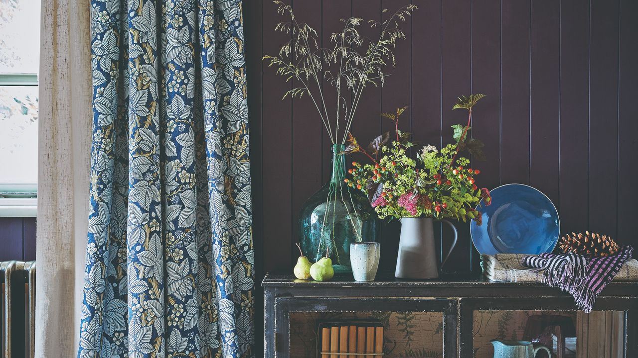 A plum-painted panelled hallway with floral curtains and a console table displaying vases of autumnal foliage