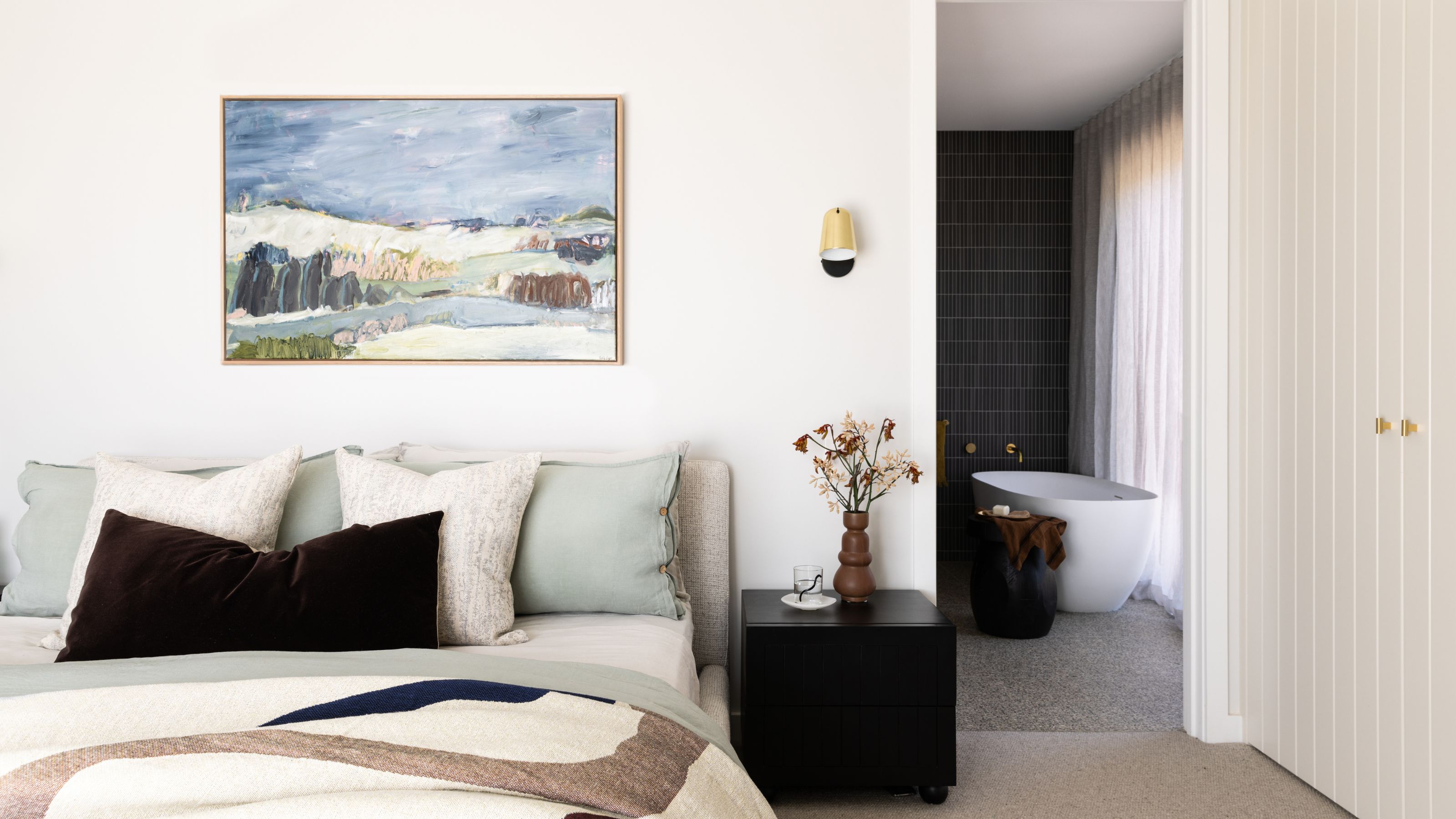 A bedroom with the view of a small ensuite bathroom. In the bathroom is a bathtub and in the bedroom there is a large bed with a painting hanging above it