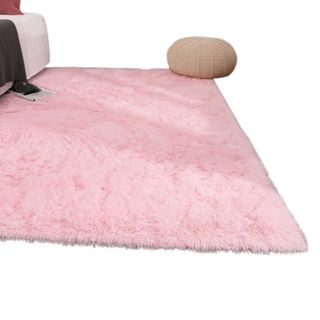 A fluffy bright pink rectangular area rug with a beige pouffle on the right corner of it and a bed on the left corner