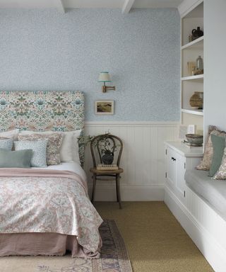 White painted bedroom with white wooden paneling,with ditsy blue floral wallpaper, floral upholstered headboard, white linen with pink and blue floral cushions, pink floral throw, natural carpet with pink and purple patterned rug, wooden chair beside bed, small painting mounted above with wall light, bespoke cabinet, bench and shelving unit in white