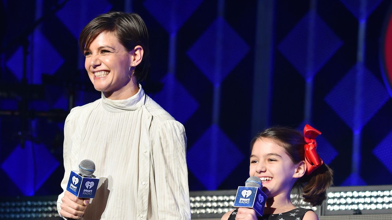 Katie Holmes smiles while wearing a floaty white shirt and holding hands with daughter Suri Holmes, who is wearing a navy dress covered in white stars