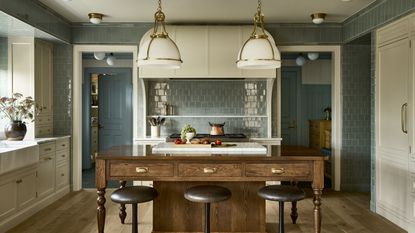 A large kitchen with light blue wall tiles, warm white cabinets, a wooden kitchen island with bar stools, and rustic pendant lights. 