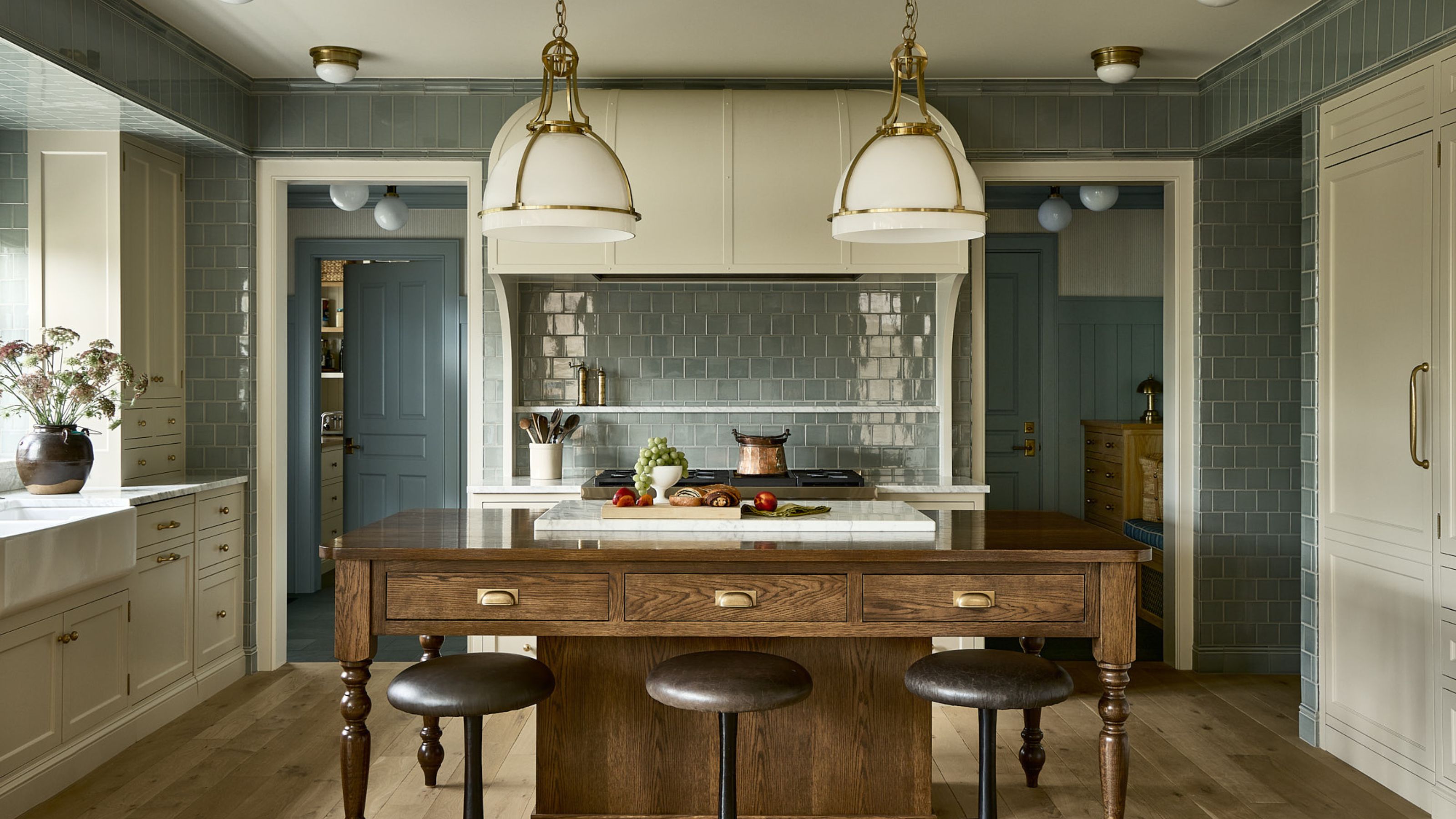 A large kitchen with light blue wall tiles, warm white cabinets, a wooden kitchen island with bar stools, and rustic pendant lights.