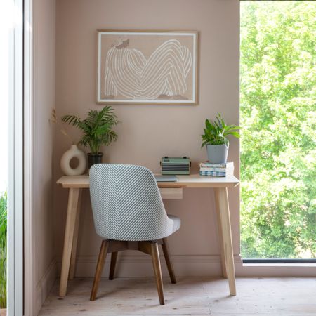 A blush pink-painted room with a small home office corner with a light wood desk and an artwork hanging above it