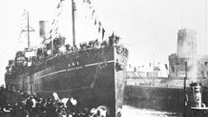 A black and white photo shows a large ship tied to a dock with people standing next to it.