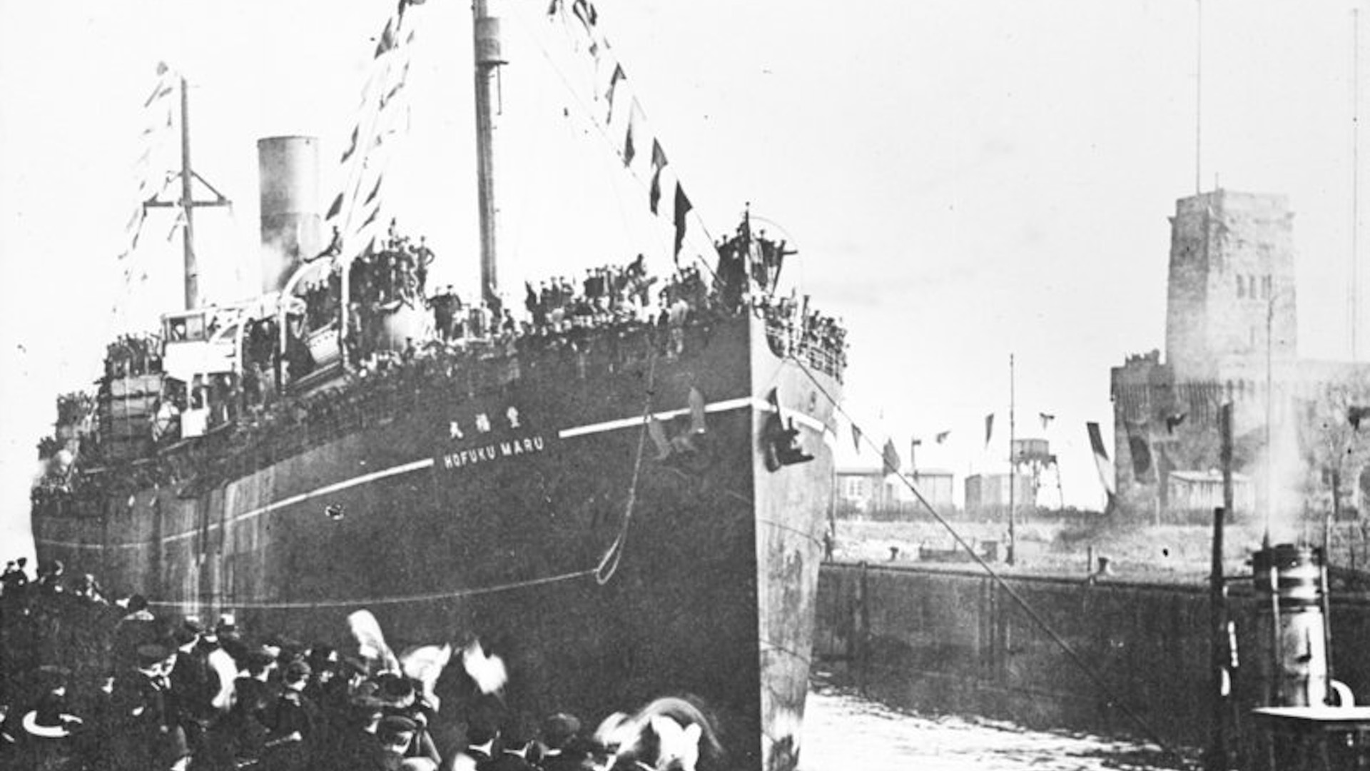 A black and white photo shows a large ship tied to a dock with people standing next to it.