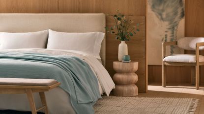 A bedroom with wood walls, an armchair, a vase of stems, a bed with a blue throw, and a bench