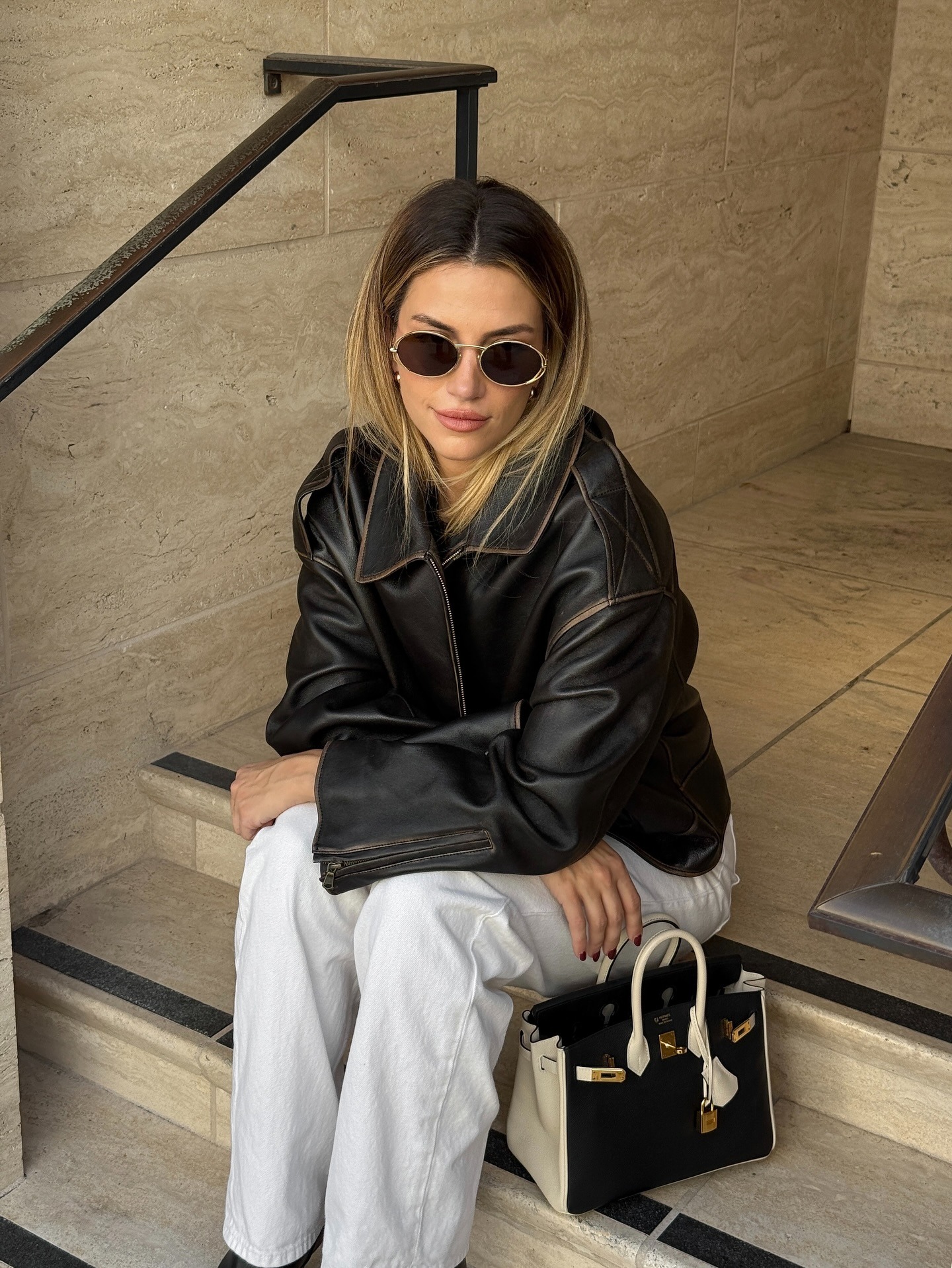 A woman sitting on a step wearing a leather jacket with dark brown hair and blonde highlights on the ends