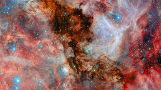 A field filled with stars and covered by clouds of gas and dust. In the centre, a thick column of dark black dust blocks light from stars that light it up from behind. More clouds behind those stars are illuminated in pale colours. Complex, layered filaments of red dust lie to the left and right. Blue, white and gold stars in various sizes can be seen around, within and through the colourful layers of dust.