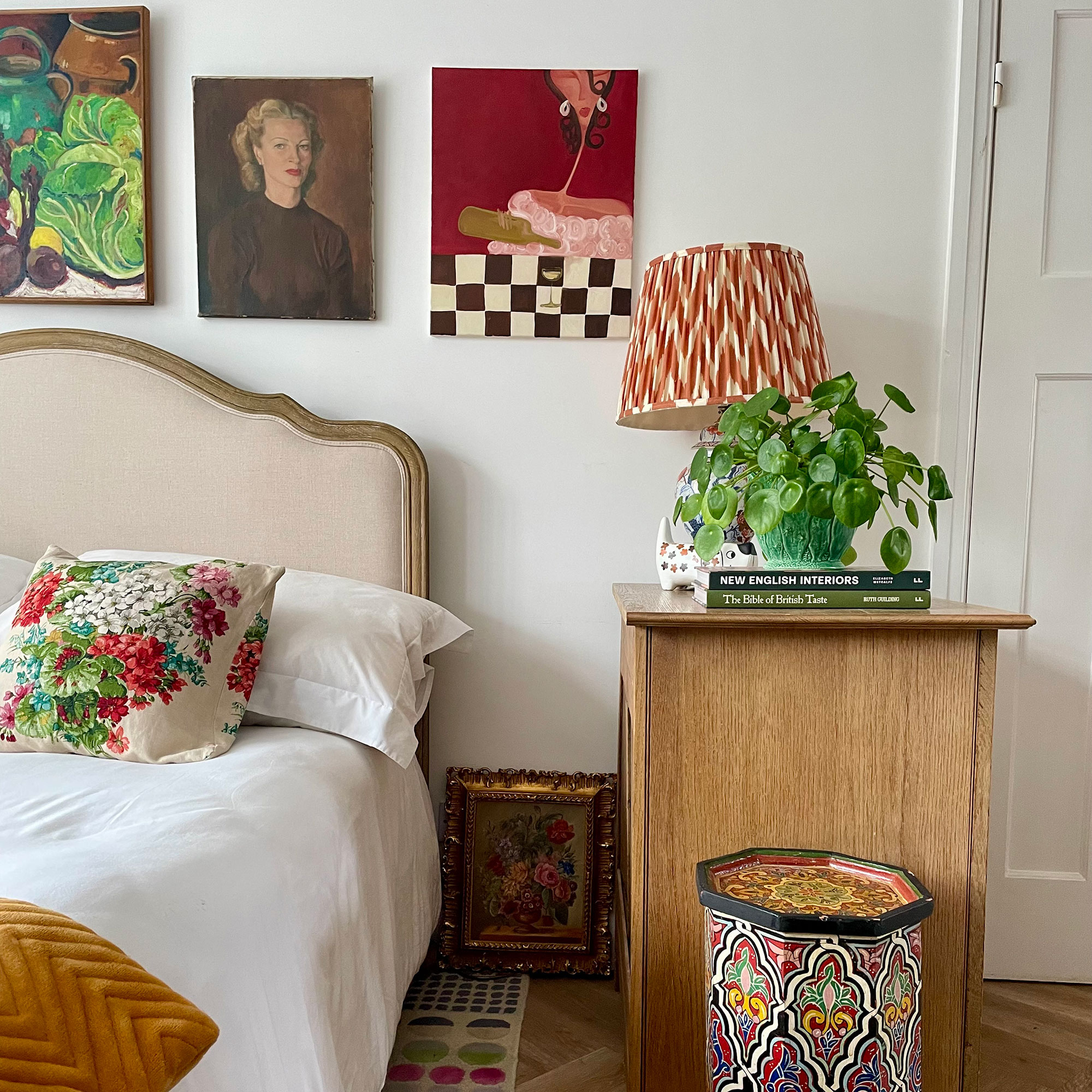 Bed with upholstered headboard in front of gallery wall and beside tall wooden table with lamp, books and plant