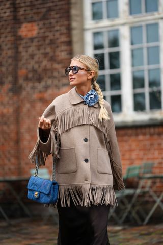 A guest wears black and blue sunglasses, a blue denim flower pattern necklace, a beige suede fringed oversized coat, a blue denim handbag from Chanel, dark brown long silk skirt, outside Paolina Russo, during the Copenhagen Fashion Week Spring/Summer 2024 on August 09, 2023 in Copenhagen, Denmark.