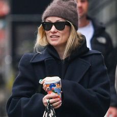 11/25/2025: Olivia Wilde is pictured on a stroll with a male companion in New York City. The American actress wore a wool beanie, black coat, checkered trousers, and Adidas trainers.