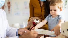a mom and doctor wearing blue face masks chat near an infant boy who's being examined at doctor's appointment. he's seated next to a teddy bear