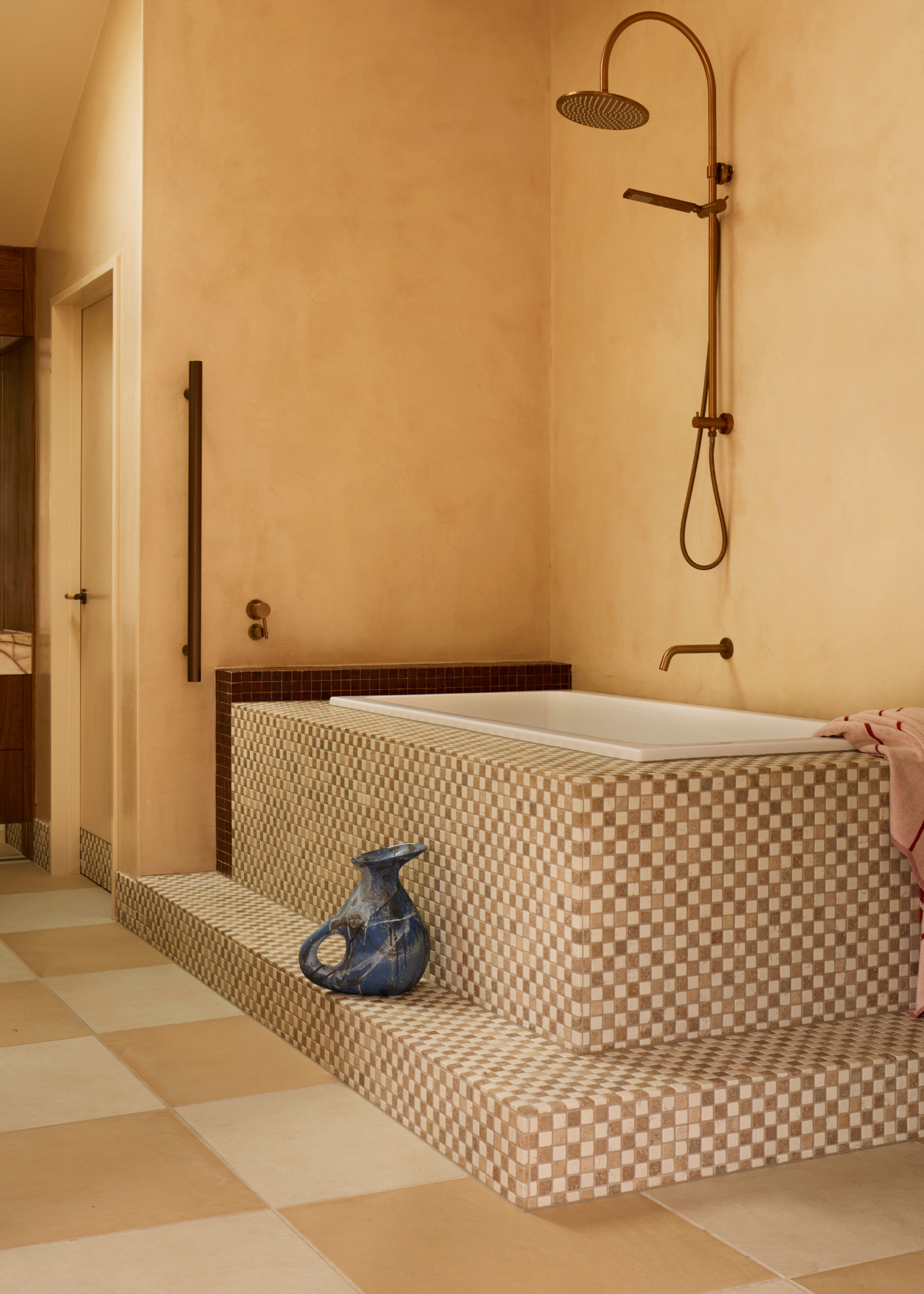 A modern bathroom in warm neutral tones with a pale-hued mosaic checkered tiled step-up bath with a gold wall-hung shower fitting and a blue jug sitting on the step