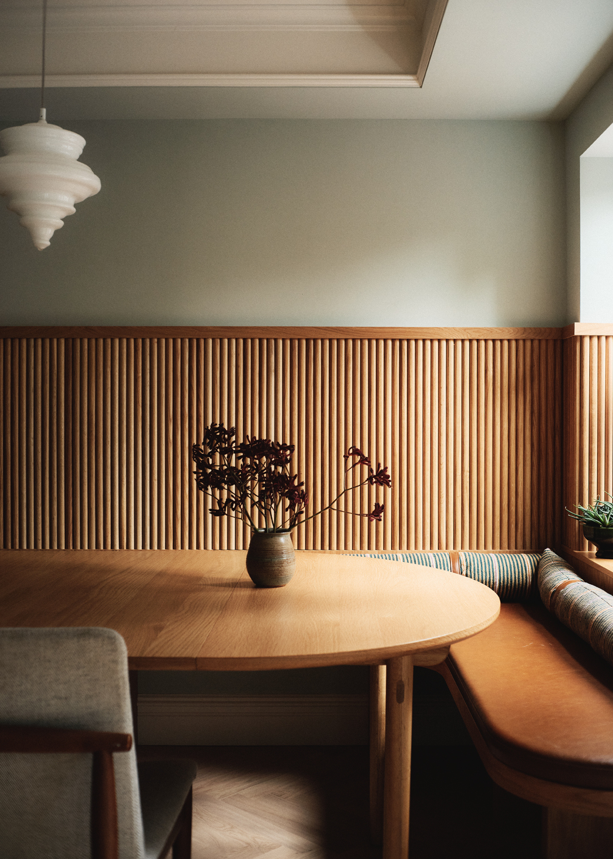 A banquette dining room with a wooden dining table featuring a ceramic vase of stems, and a tiered crystal light above